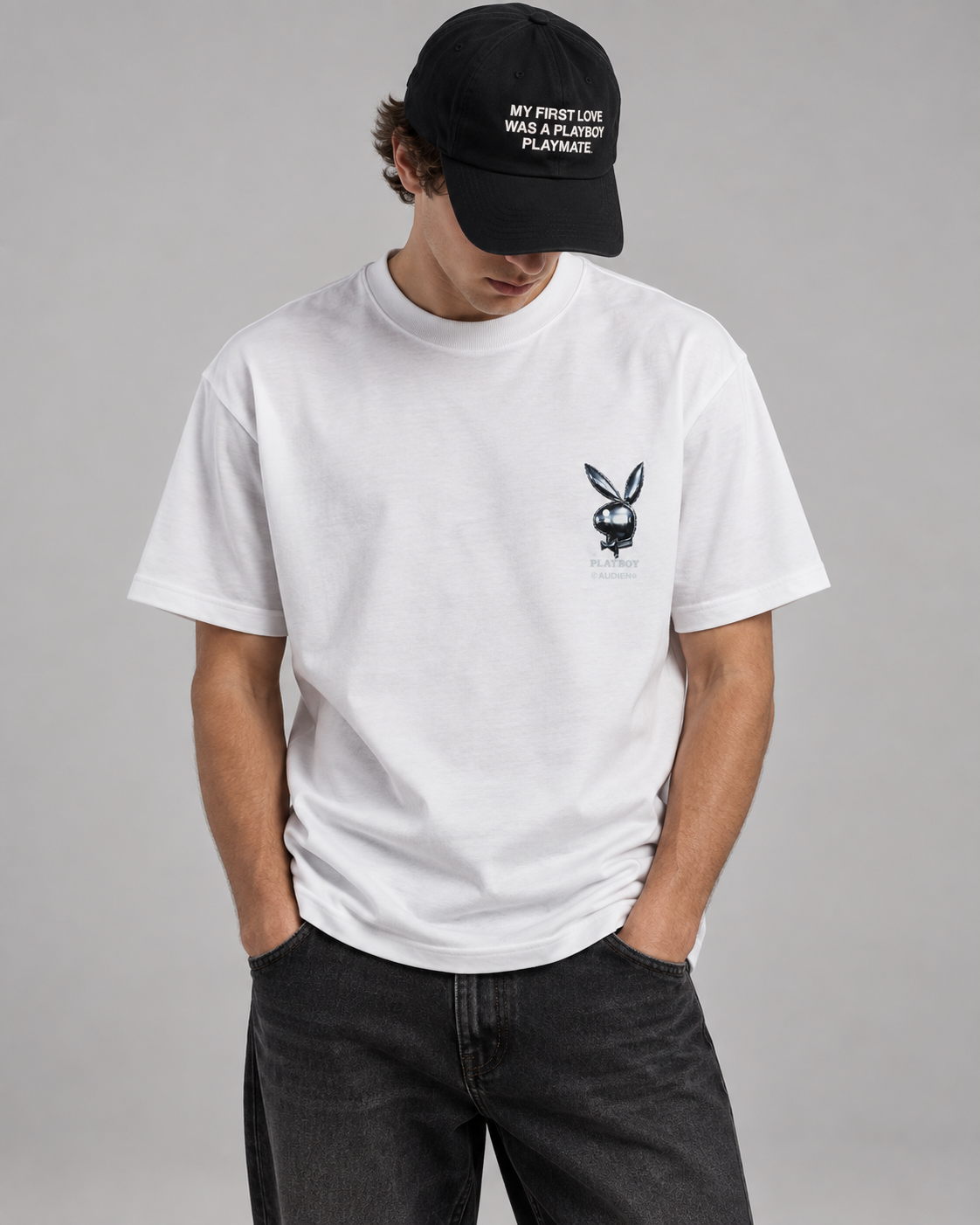 A man wearing a white short-sleeve crewneck t-shirt with a small black Playboy bunny logo on the left chest, paired with a black baseball cap and dark gray jeans, against a solid gray studio background.