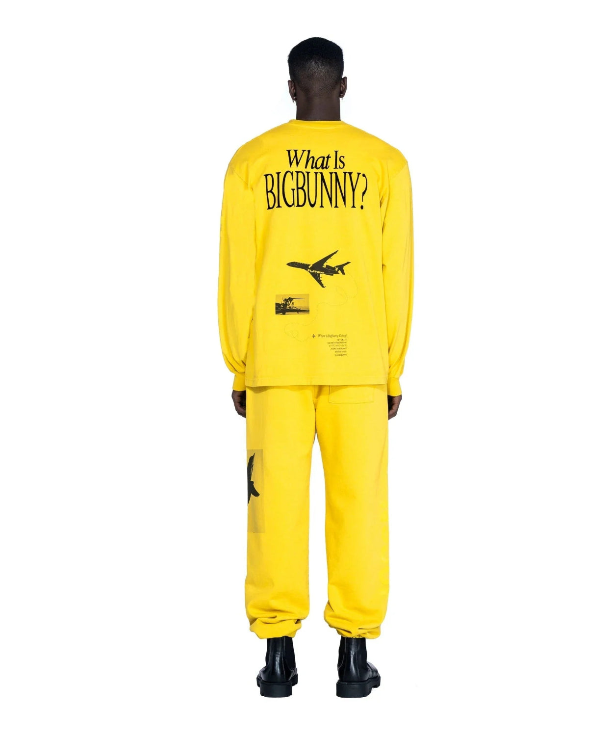 Man wearing bright yellow long sleeve shirt and pants set with airplane and 'What Is BIGBUNNY?' text on back, viewed from behind.