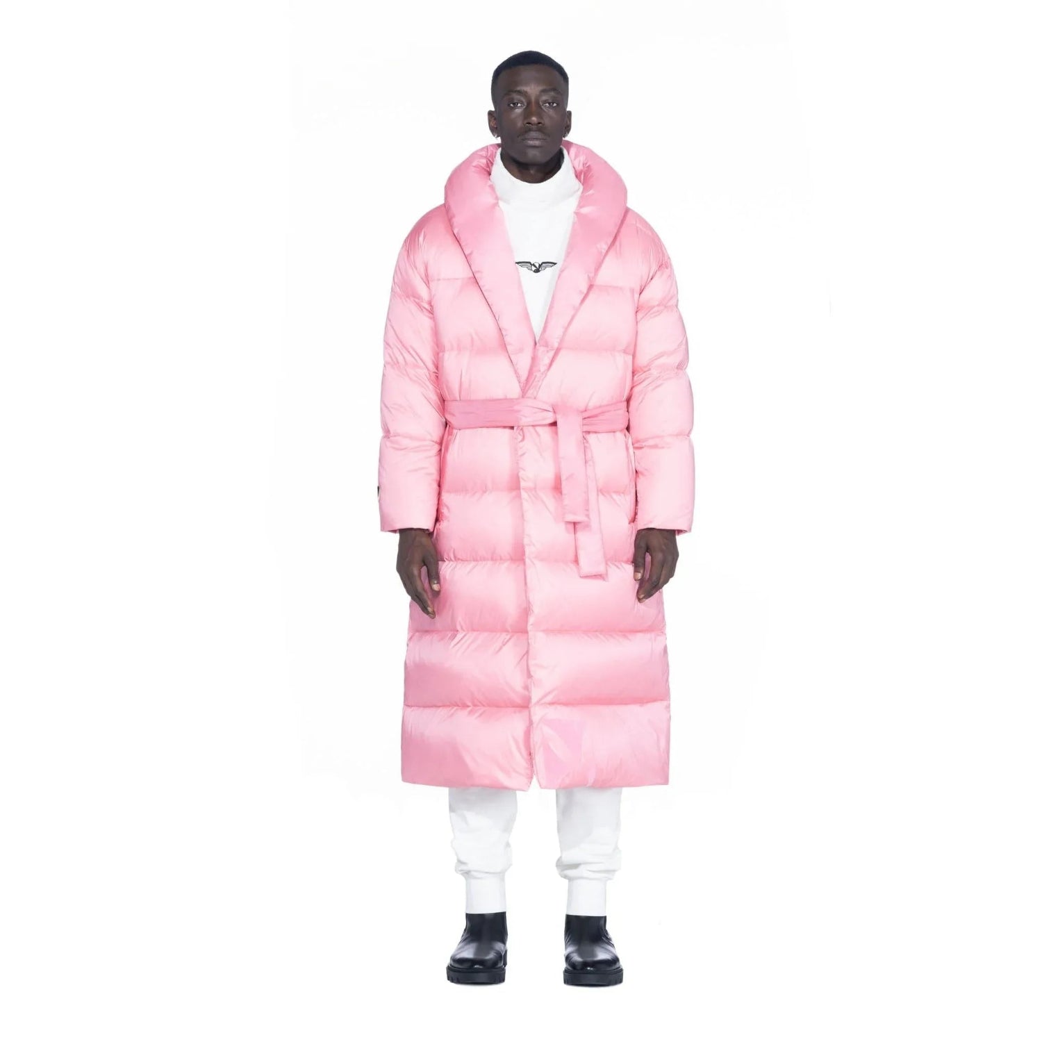 Man wearing a pink belted long puffer jacket and white pants, standing against white background