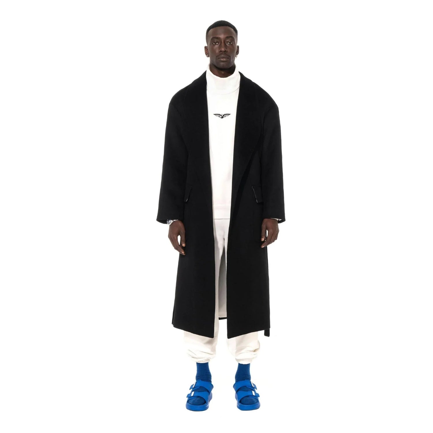 Man wearing long black wool robe coat, white turtleneck, white pants, and blue sandals