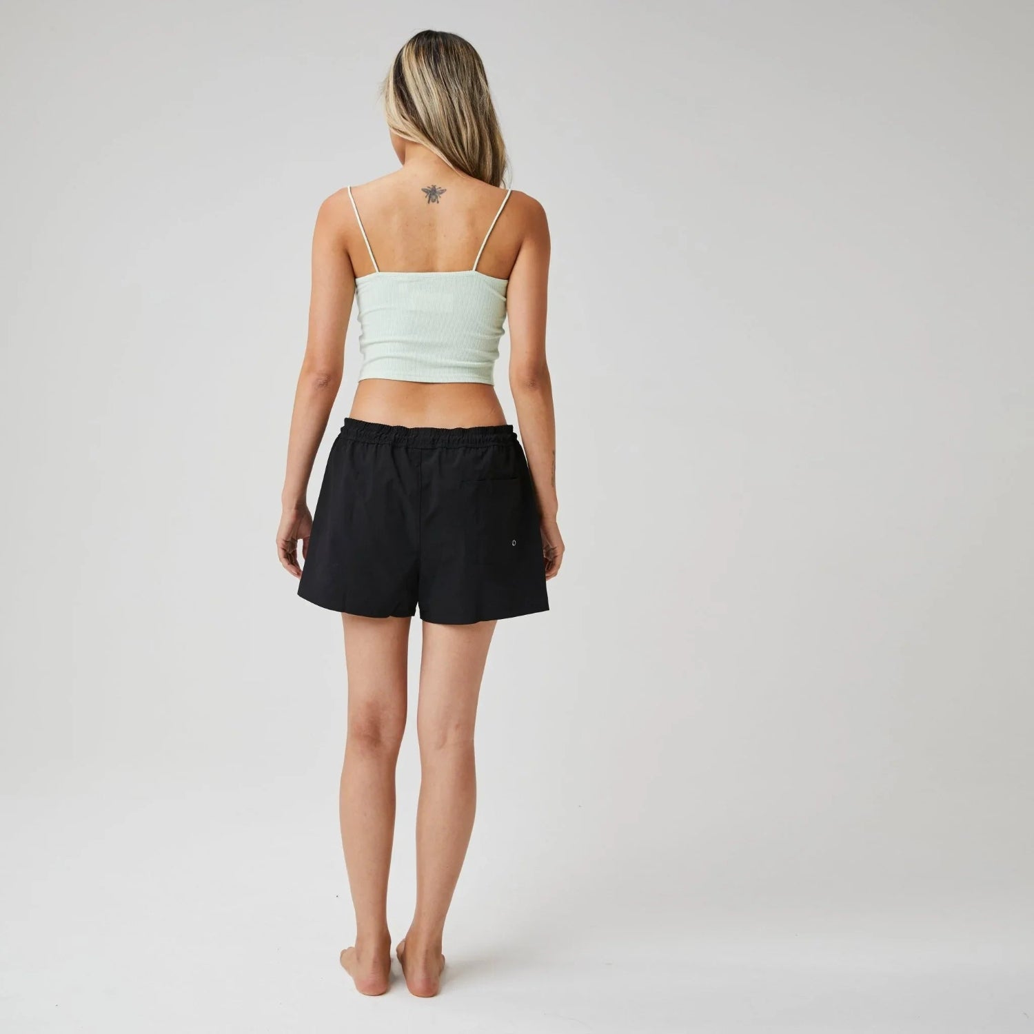 Woman wearing black board shorts and mint green tank top, back view, studio setting