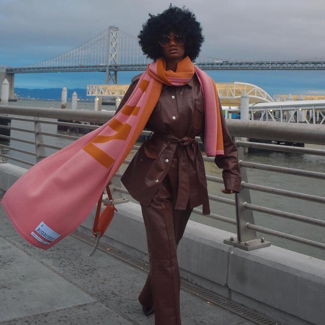 Woman wearing crocodile leather flare pants and jacket, pink scarf, by waterfront bridge