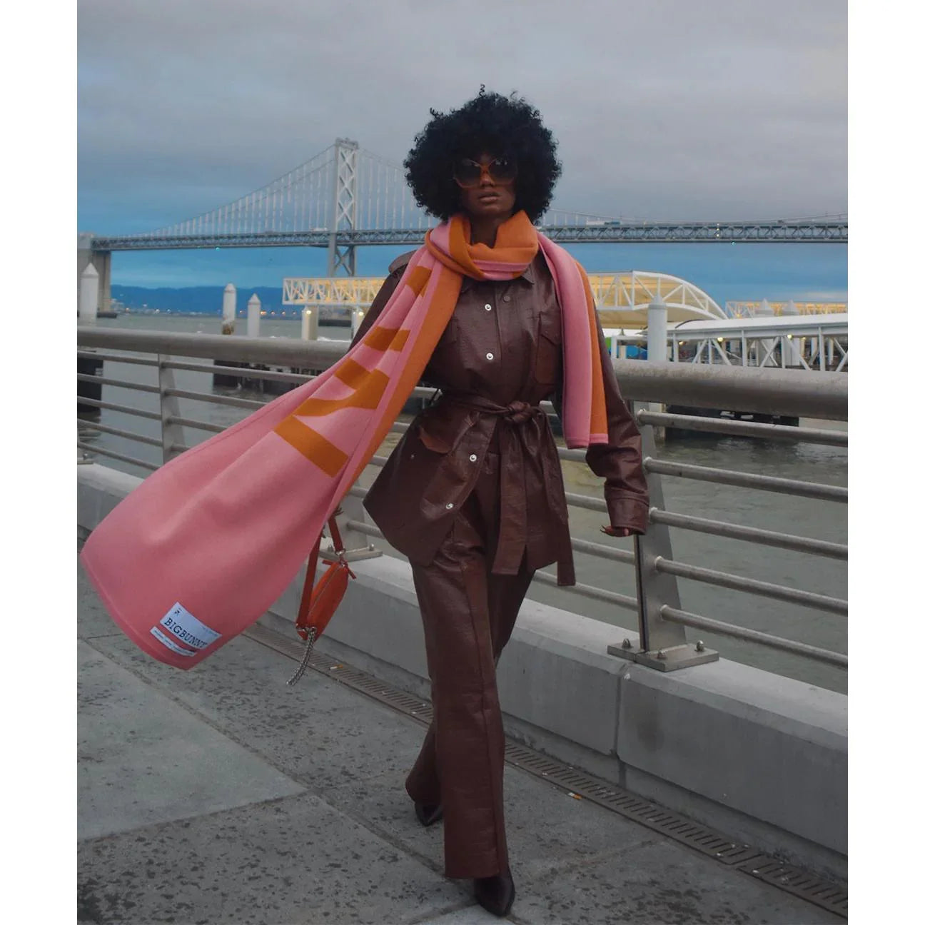 Woman in brown crocodile leather safari jacket and pants with pink orange scarf, outdoor city bridge scene
