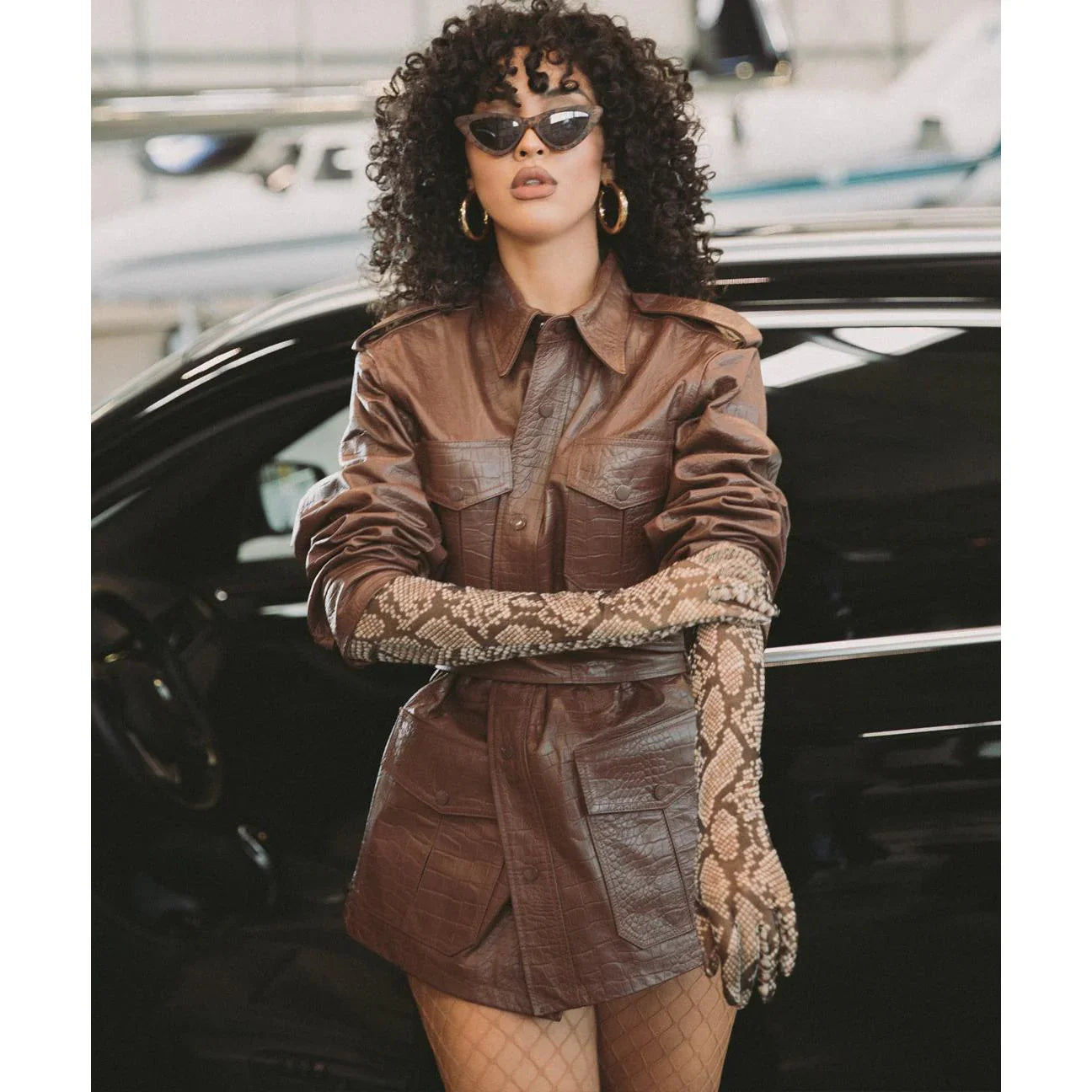 Woman in brown crocodile leather safari jacket, snake print gloves, and sunglasses standing by a car.