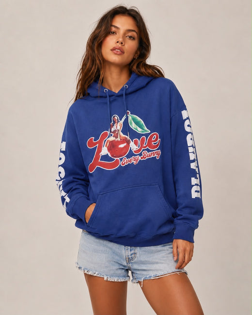 A woman modeling a royal blue hoodie with a colorful cherry graphic on the front and white text down the sleeves, paired with light-wash denim shorts against a plain beige studio background.