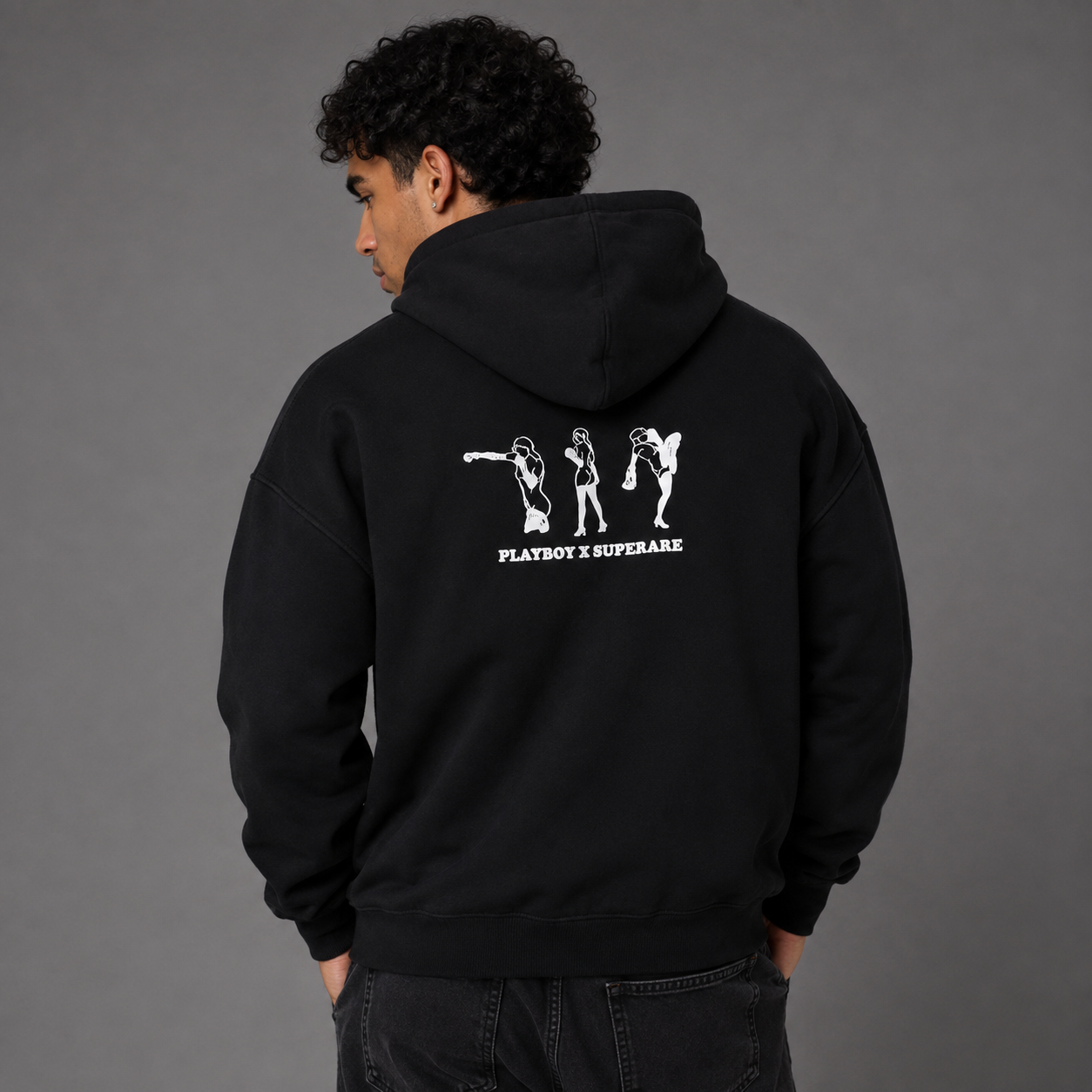 A man wearing a black hoodie from the back, featuring a white graphic print of three dancing figures and the text 'PLAYBOY X SUPERSTAR' on the back, set against a solid gray studio background.