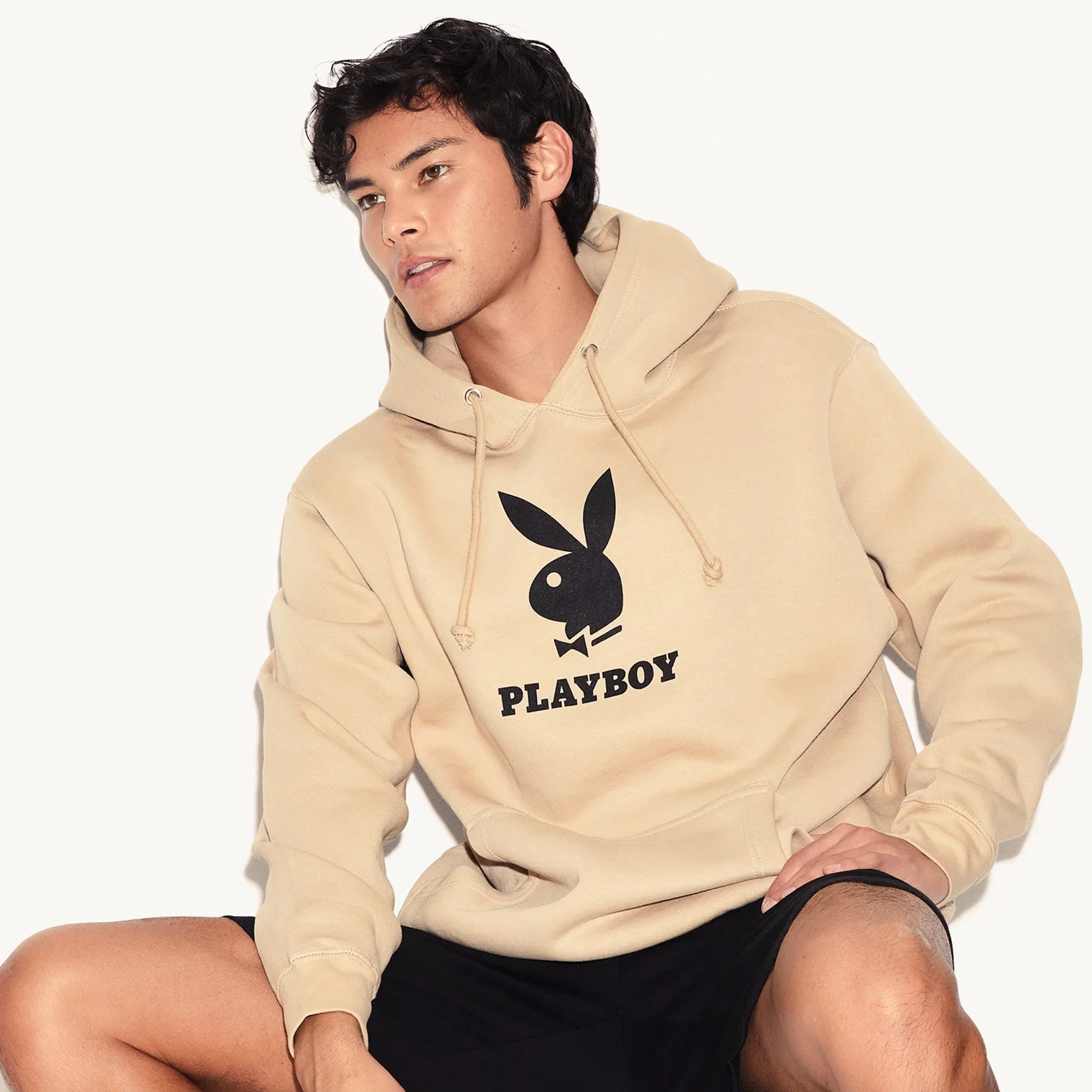 Man wearing beige hoodie with Playboy bunny logo and text, casual lifestyle fashion