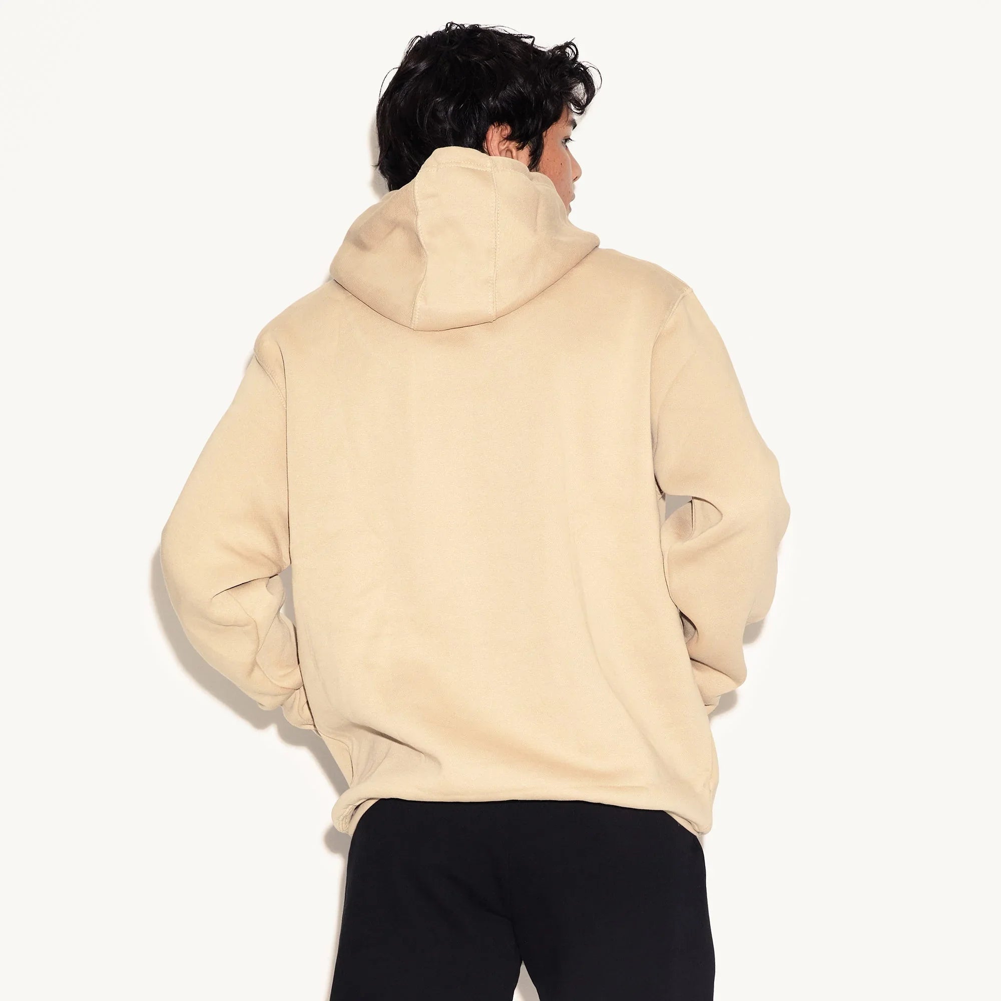 Back view of a man wearing a beige hooded sweatshirt with black pants on a plain background.