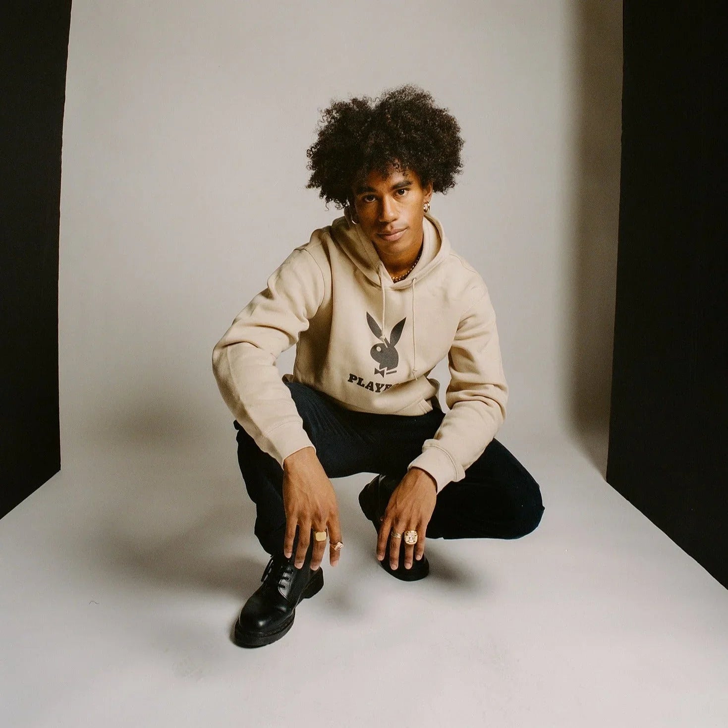Man wearing beige hoodie with bunny logo, black pants, black shoes, studio background
