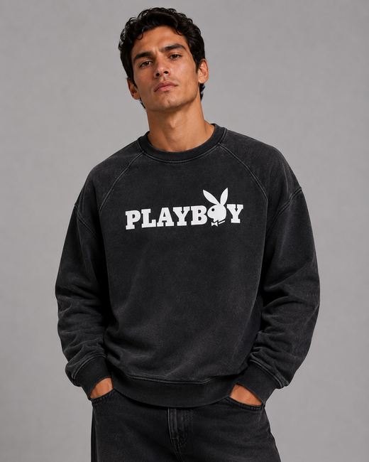 A man wearing a black washed sweatshirt with a white PLAYBOY logo and bunny graphic on the chest, paired with dark jeans, against a solid gray studio background.