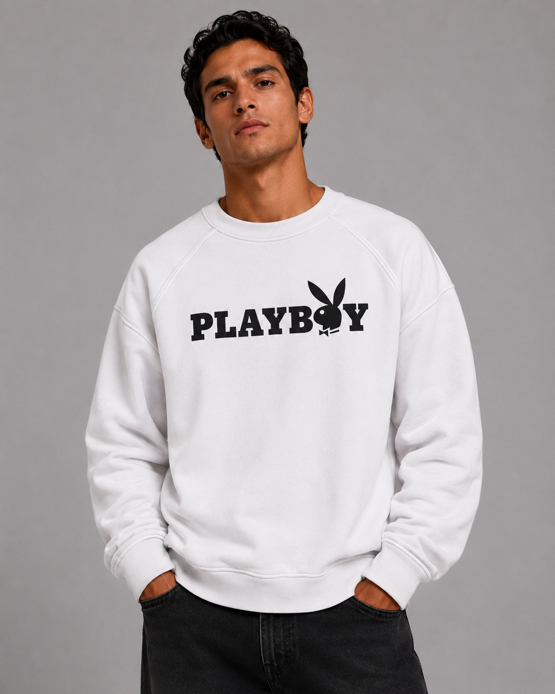 A man wearing a white crewneck sweatshirt with a black PLAYBOY logo across the chest, paired with dark jeans, photographed against a solid gray studio background.