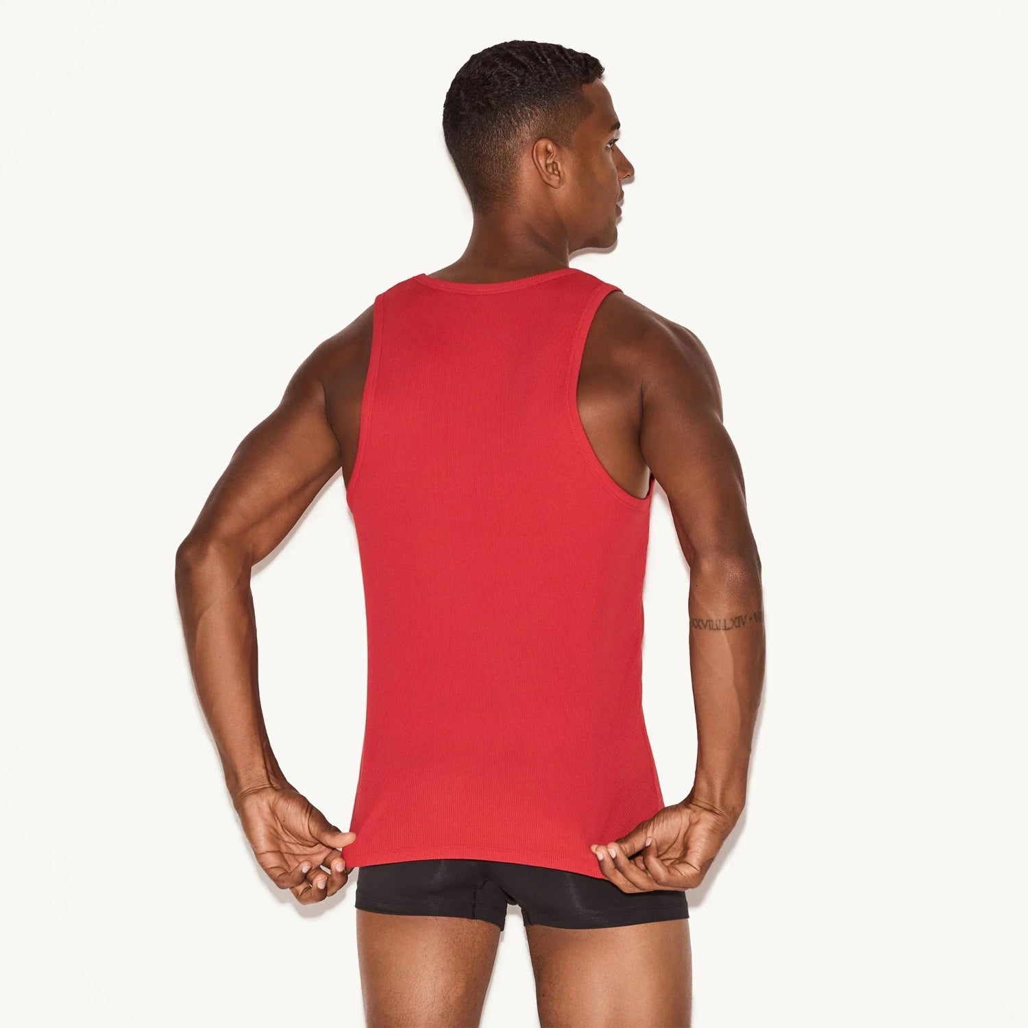 Man wearing red ribbed tank top and black shorts, shown from the back, on white background