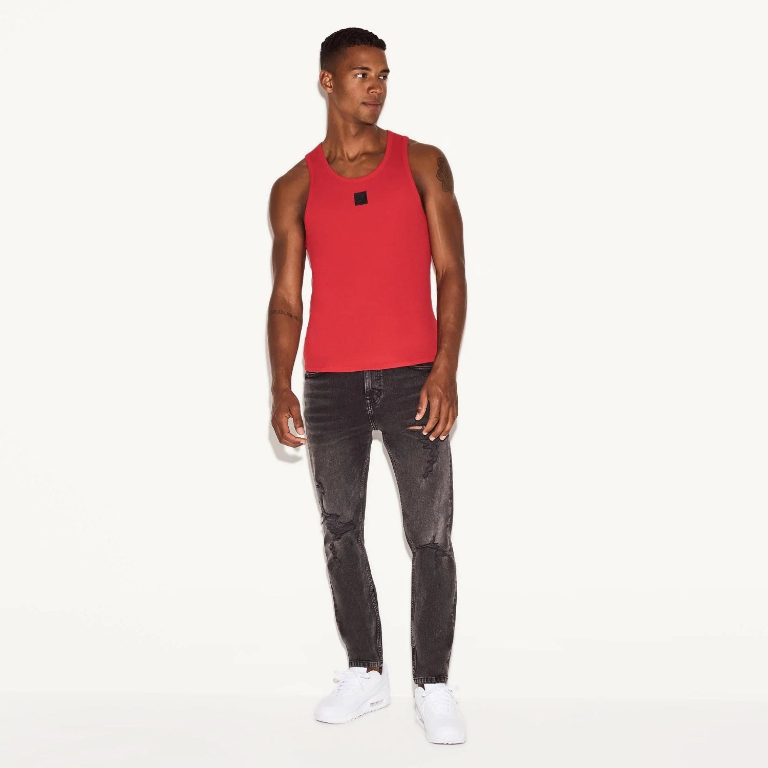 Man wearing a red ribbed tank top, black jeans, and white sneakers, standing against a white background.