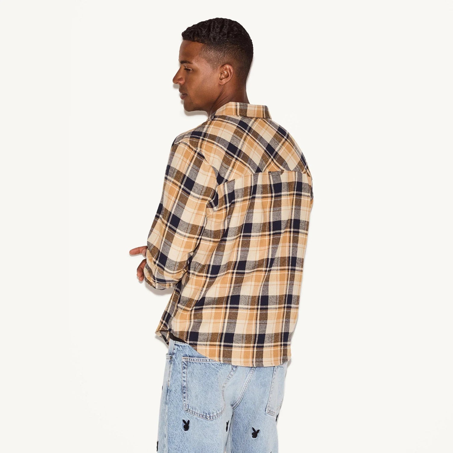 Man wearing tan plaid flannel shirt and light wash jeans with embroidered logos, side view
