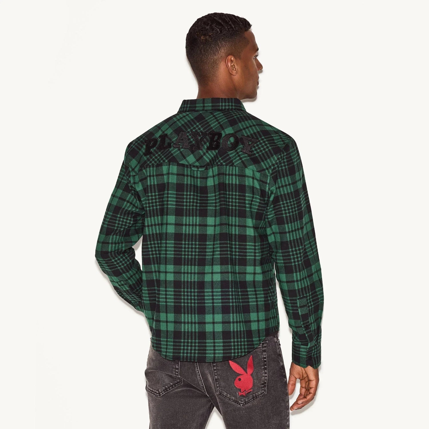 Man wearing green plaid flannel long sleeve shirt and black jeans with red bunny patch