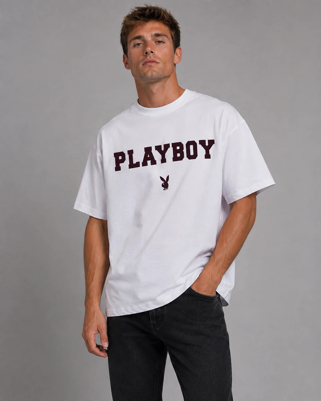 A man wearing a white Playboy t-shirt with maroon collegiate lettering, against a grey studio background.