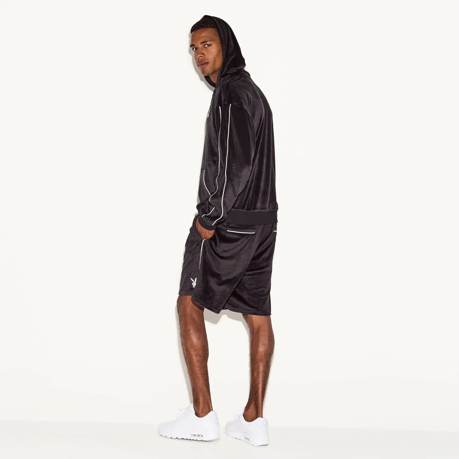Man wearing black velour hoodie and shorts set with white piping, side view