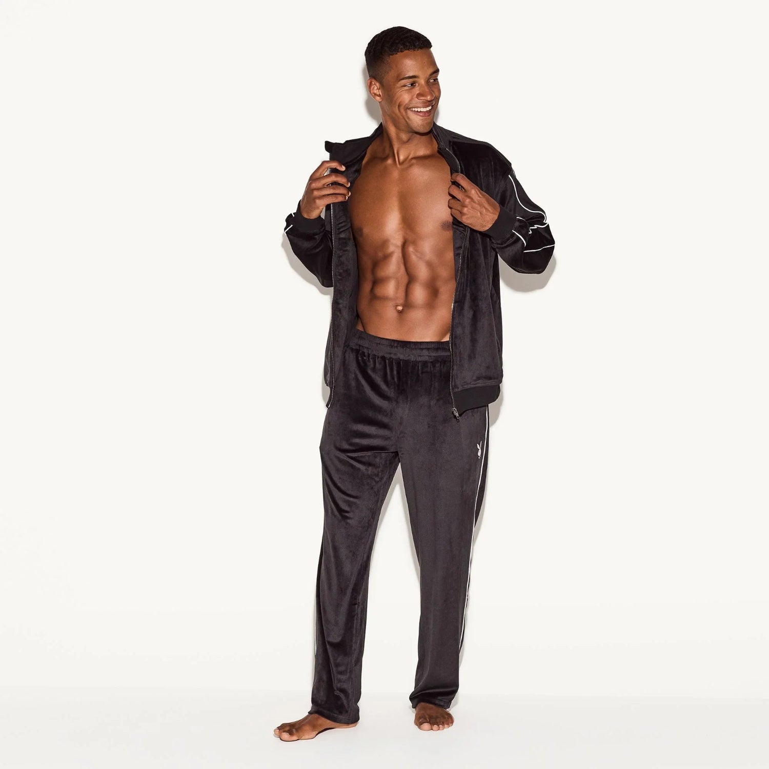 Man wearing black velour track pants and matching jacket with white piping details