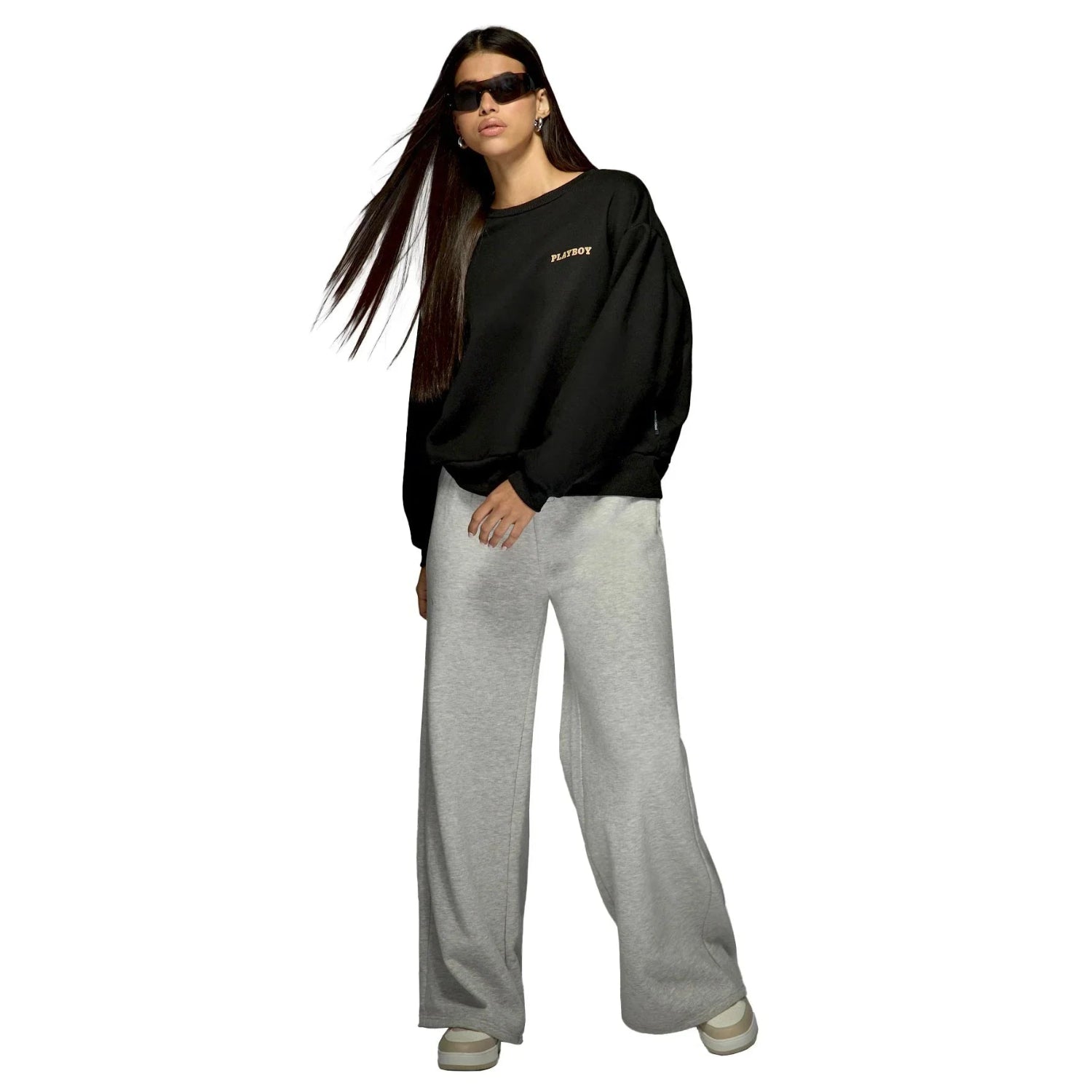 Woman wearing black sweatshirt with small logo and gray wide-leg sweatpants, standing against white background