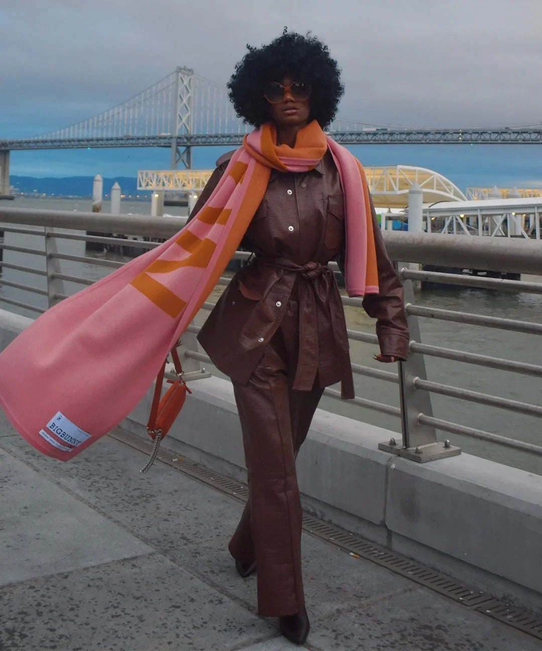 Stylish person in brown leather outfit with pink and orange cashmere scarf outdoors by bridge