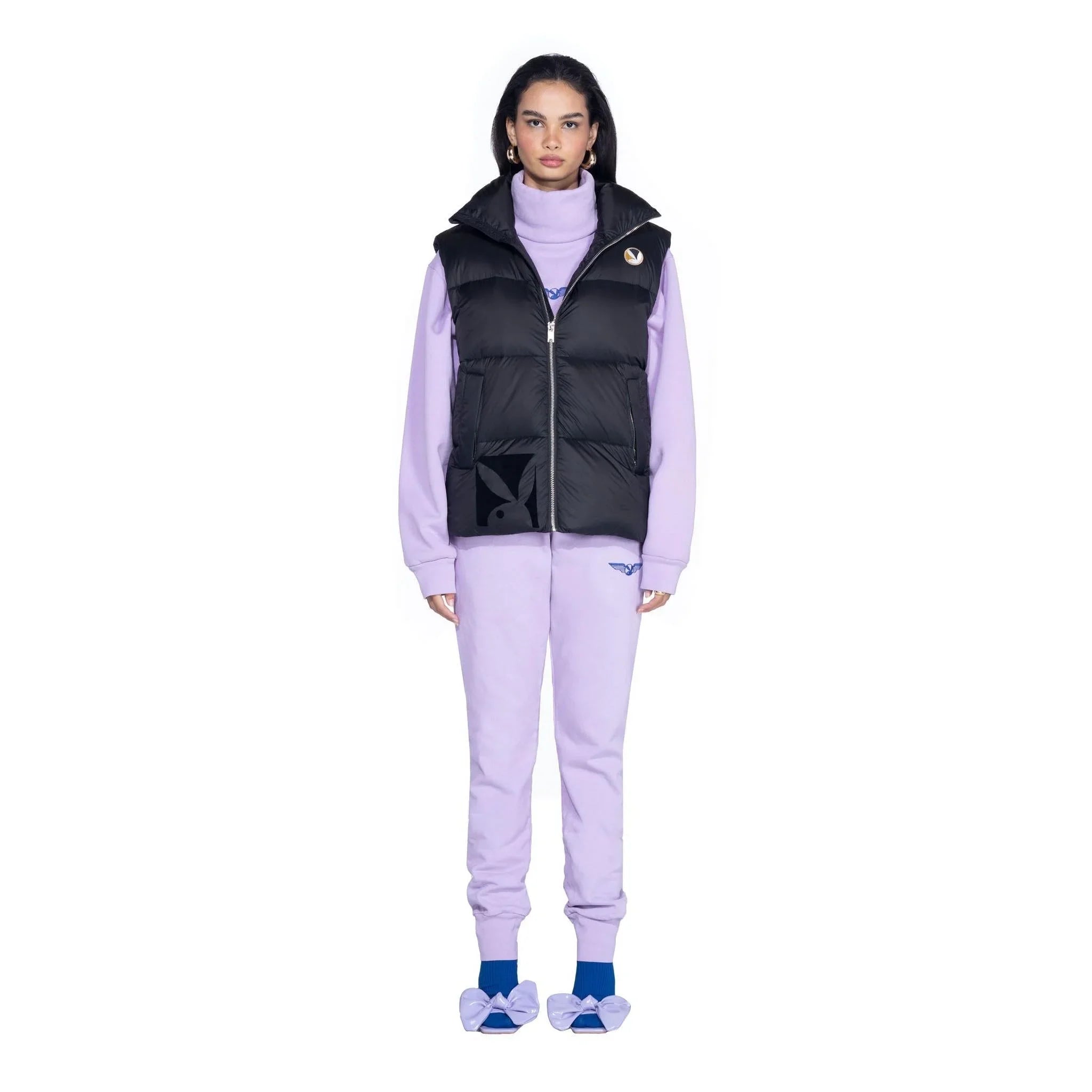 Woman wearing a black puffer vest over a lavender tracksuit with blue bow slippers, studio shot.