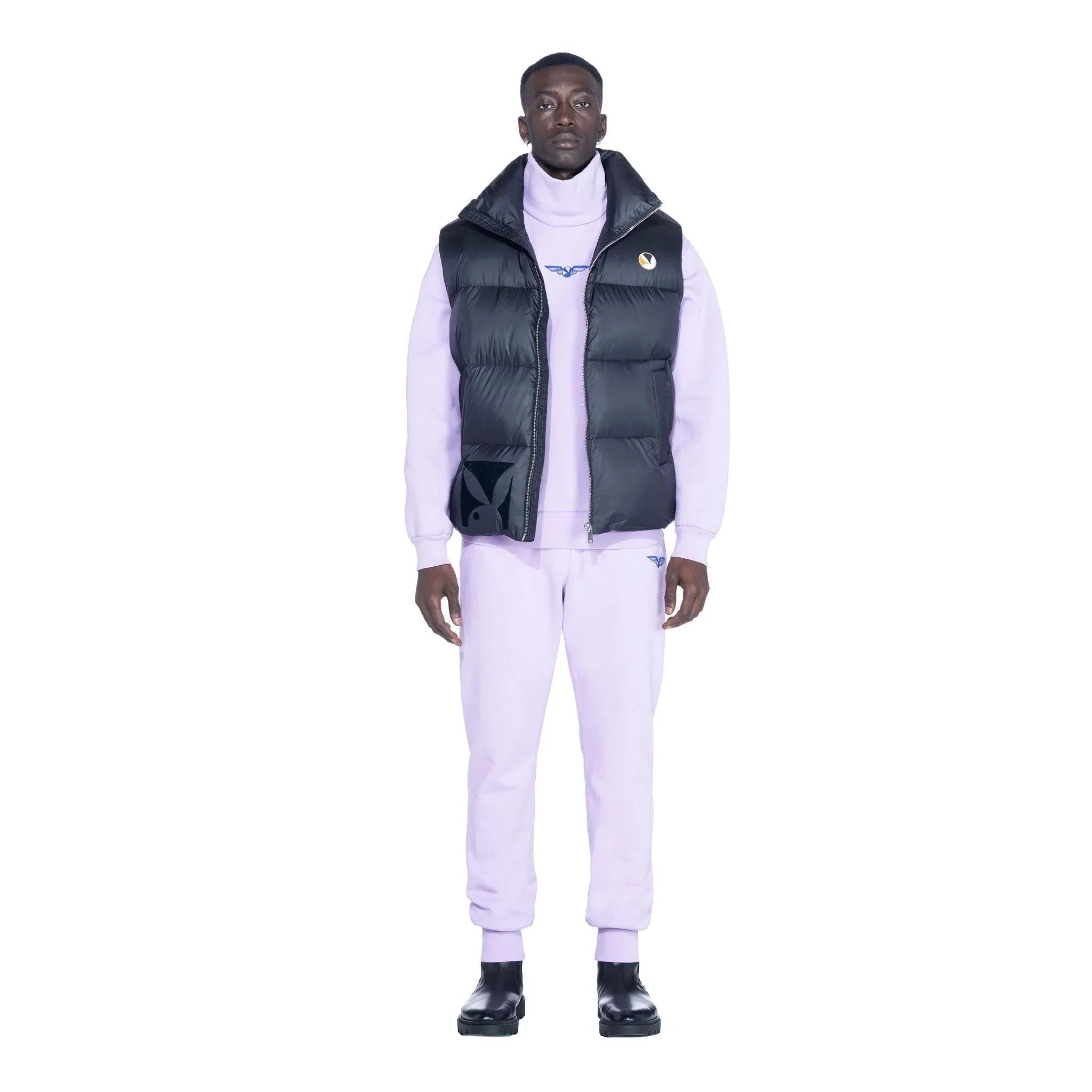 Man wearing black puffer vest over lilac tracksuit with logos, standing against white background