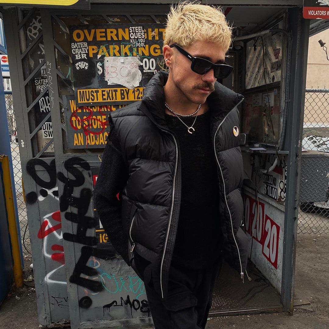 Man wearing a black puffer vest and sunglasses in an urban, graffiti-covered setting