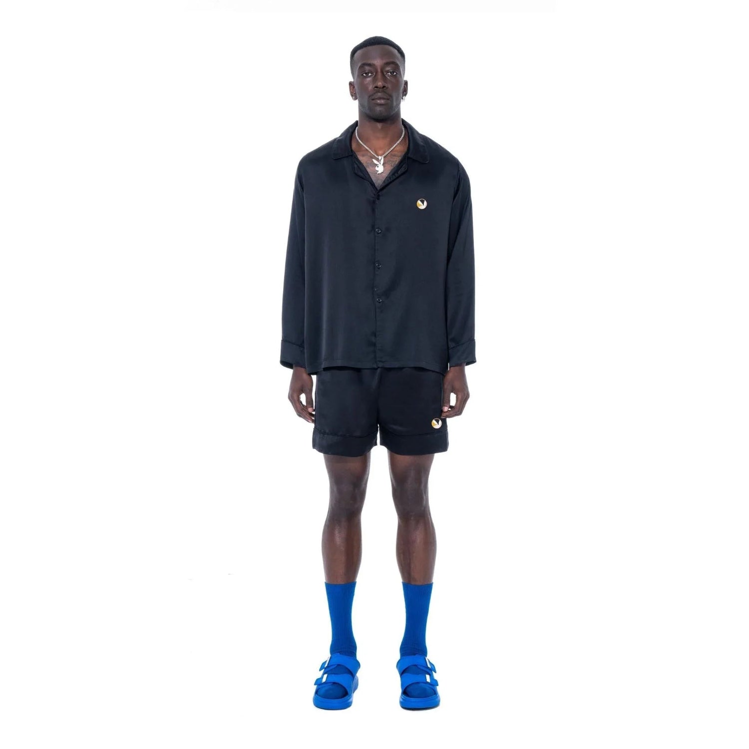 Man wearing black silk button-down collared top, black shorts, blue sandals and socks