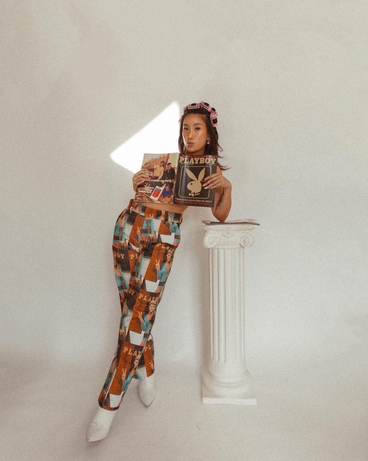 Woman in retro print pants with magazine covers, holding vintage magazines, posing by a white column