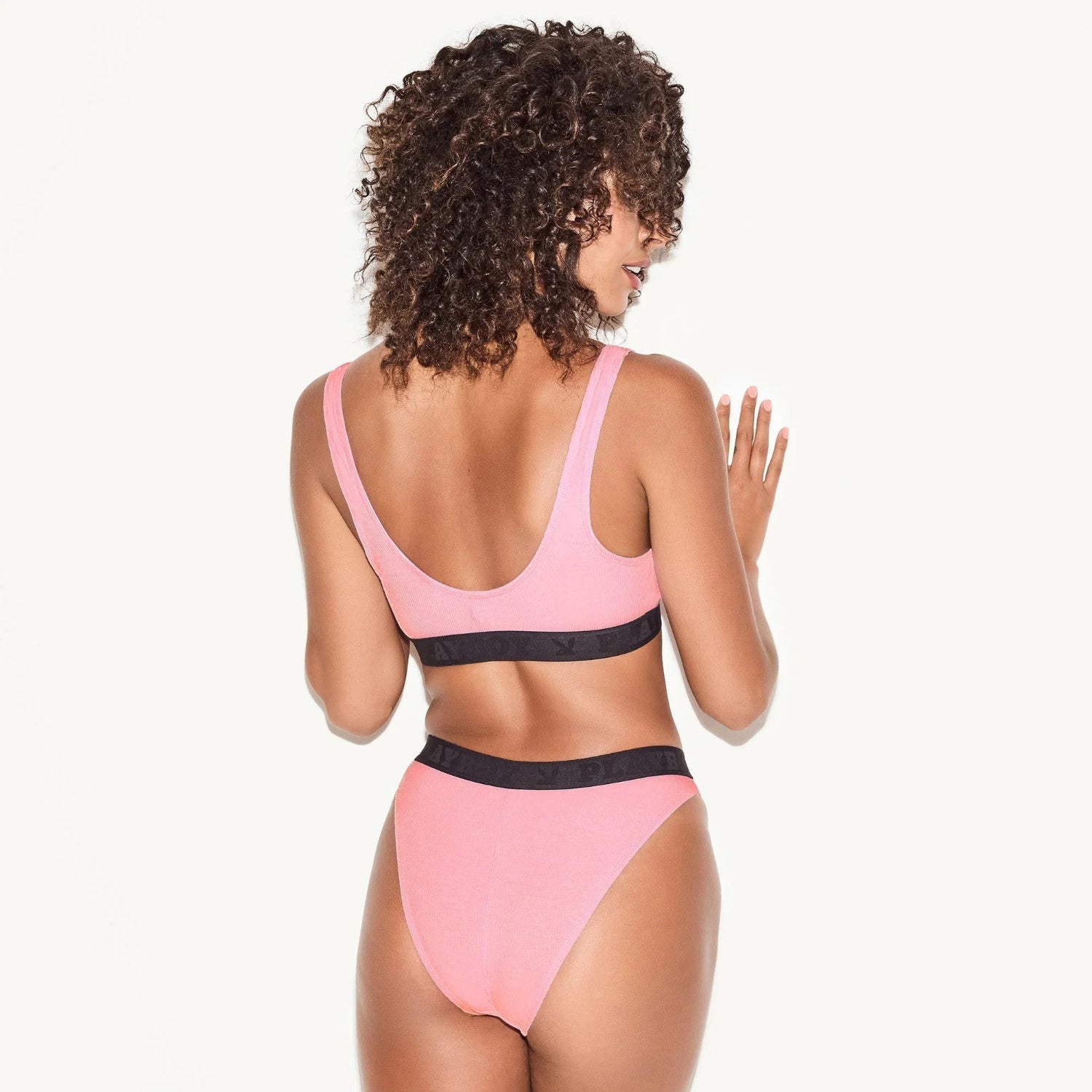 Woman wearing pink ribbed bralette and high-waisted briefs with black waistband, back view