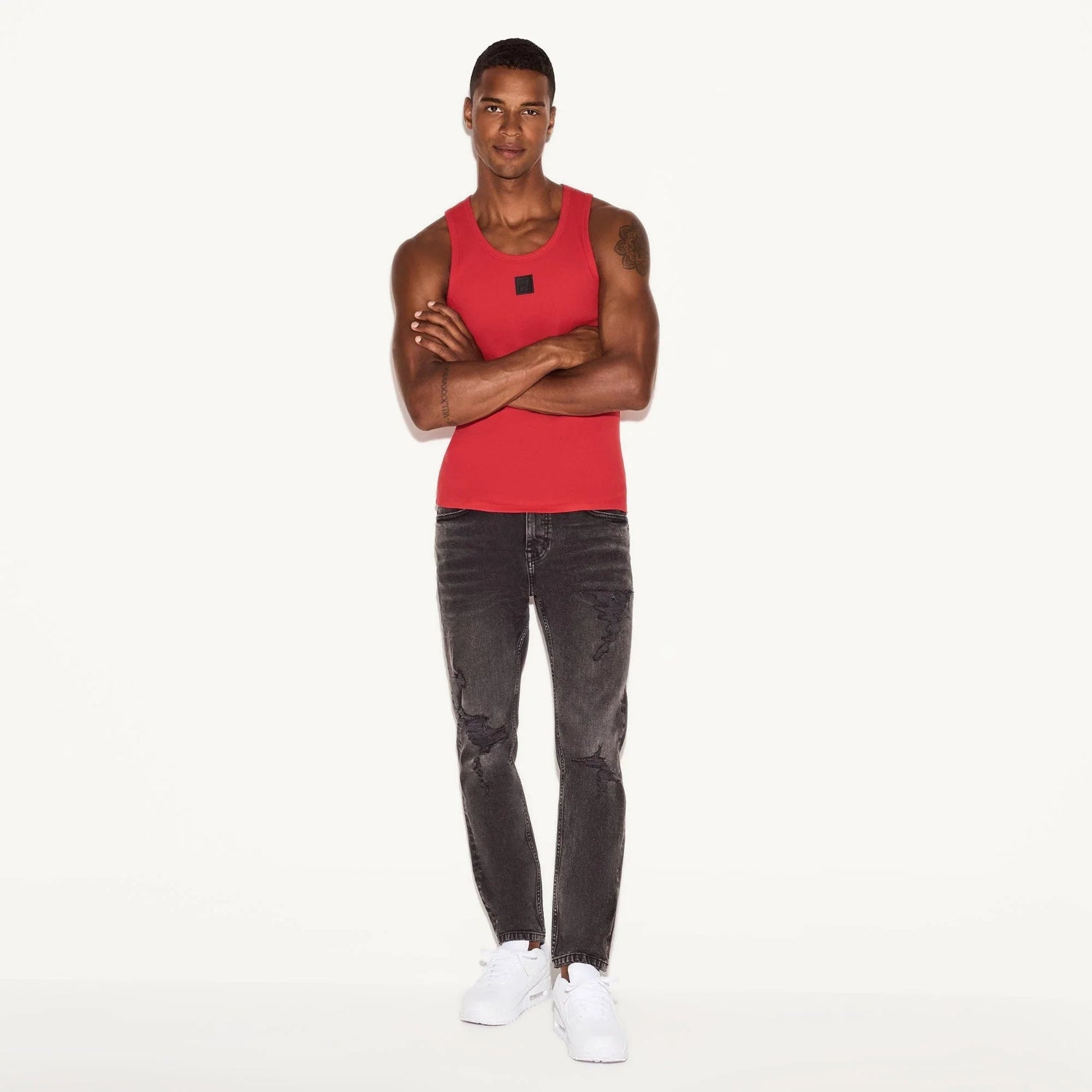 Man wearing distressed grey skinny jeans, red tank top, and white sneakers, standing casually