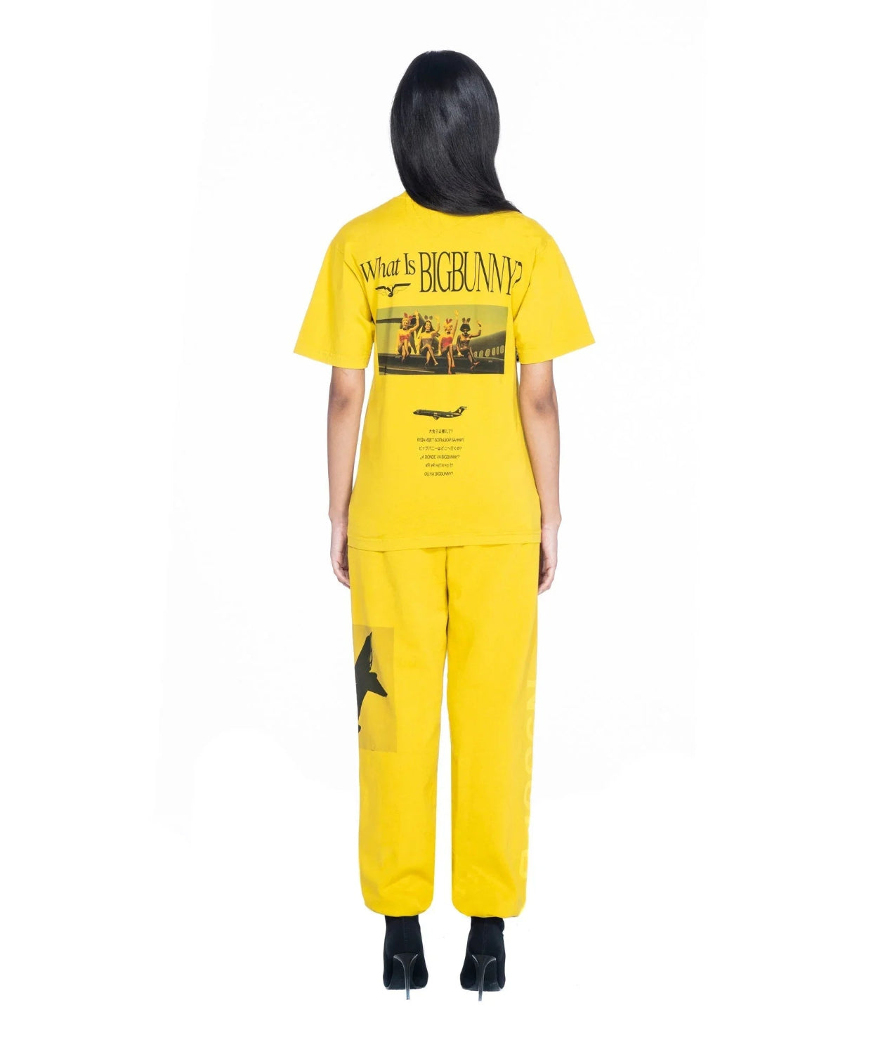 Woman wearing yellow graphic t-shirt and pants, rear view, bold text and print on back.