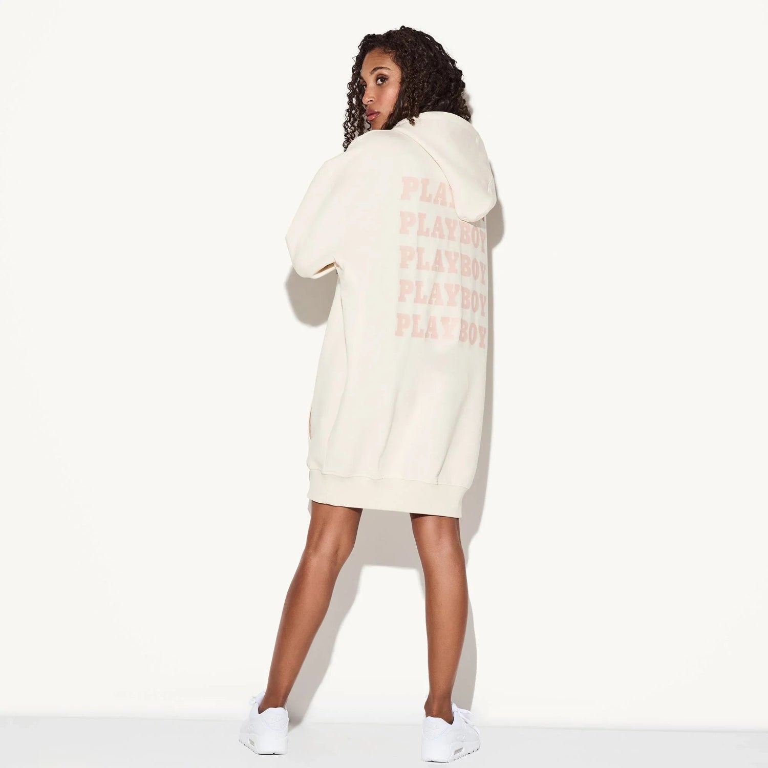 Woman wearing cream hoodie dress with repeat logo print on back, casual streetwear style