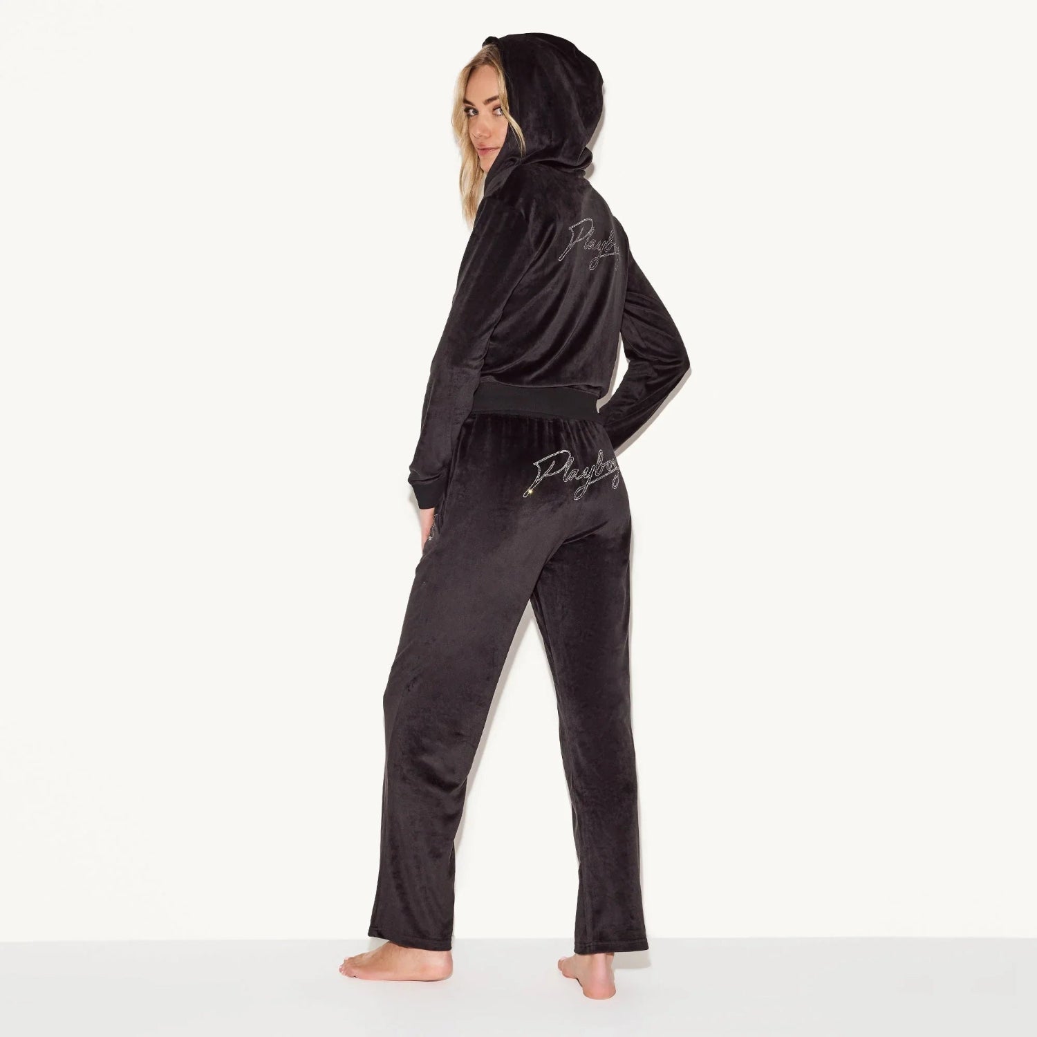 Woman wearing black velour tracksuit with script logo on back, hood up, facing sideways