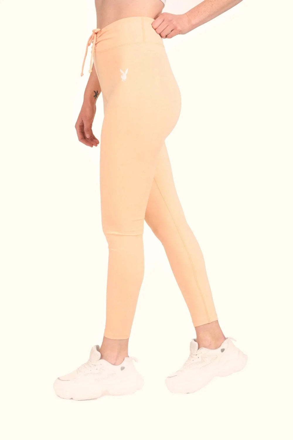 Woman wearing peach high-waisted ruched leggings with drawstring and white sneakers