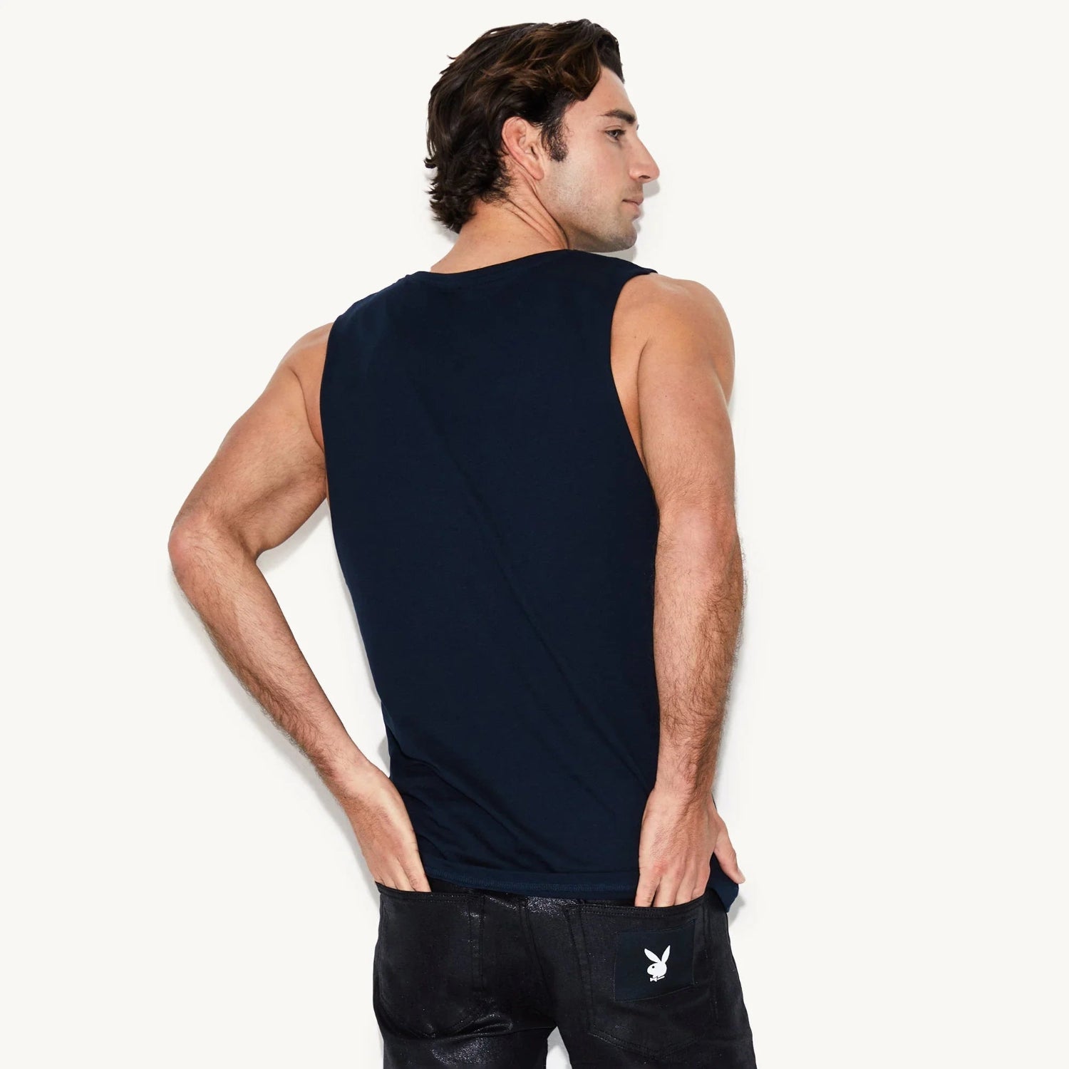 Man wearing navy muscle tank and black jeans with bunny logo patch on pocket, rear view
