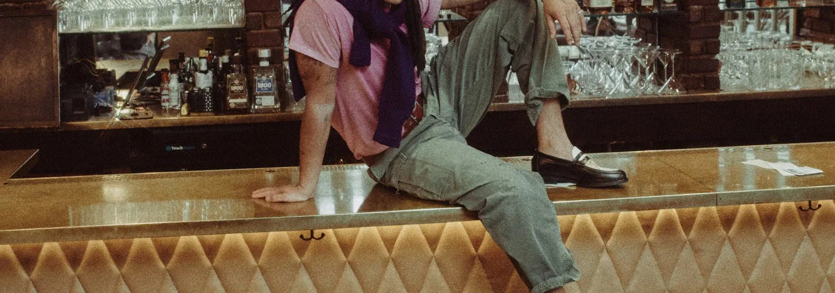 Man in casual fashion with pink t-shirt, green pants, and loafers sitting on a bar counter