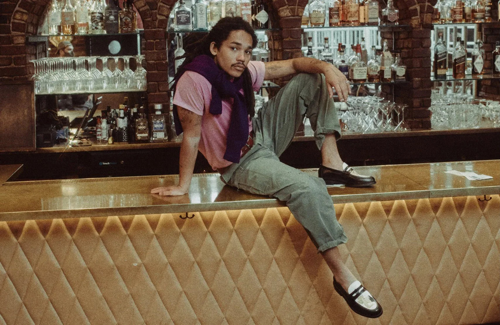 Man in casual fashion with pink t-shirt, green pants, and loafers sitting on a bar counter