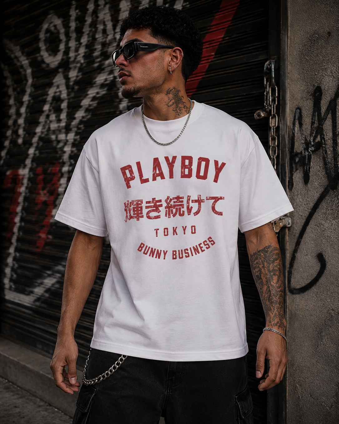 A man wearing sunglasses and a white Playboy Tokyo t-shirt with red Japanese text and bunny business graphics, standing in front of a graffiti-covered metal shutter. #Streetwearmen