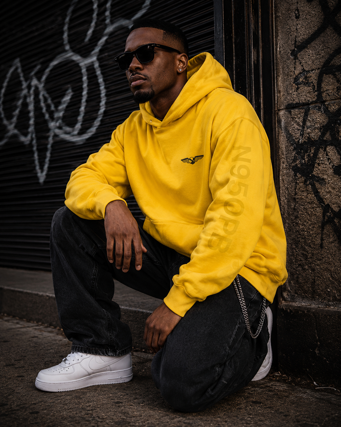 A man wearing a bright yellow hoodie with a small black bird logo on the left chest, paired with black jeans and white sneakers, crouching against a dark graffiti-covered wall. #Streetwearmen