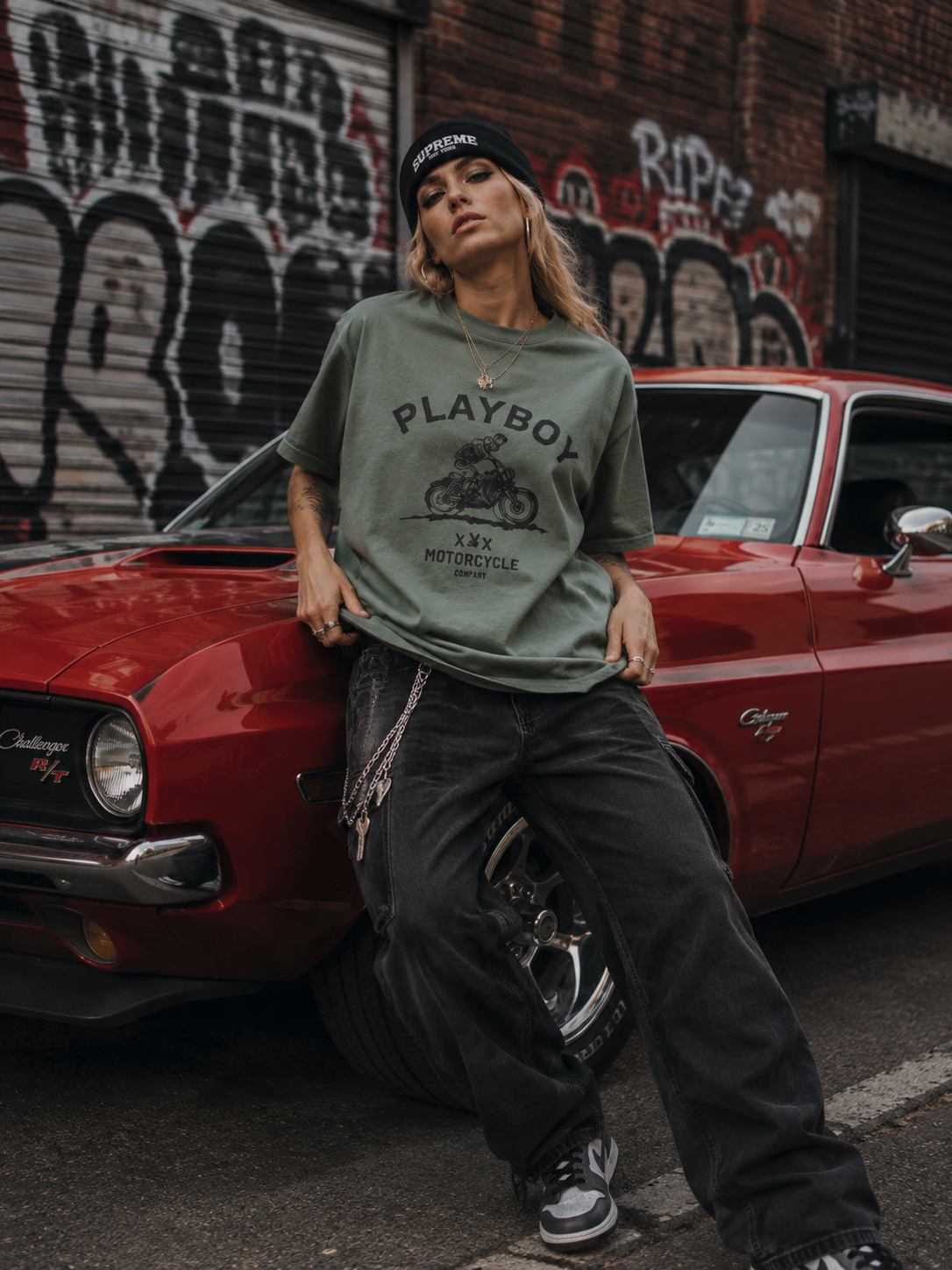 A woman posing in front of a red classic car, wearing an oversized olive green graphic t-shirt with a Playboy logo, dark baggy jeans, and a black beanie. #Streetwearwomen