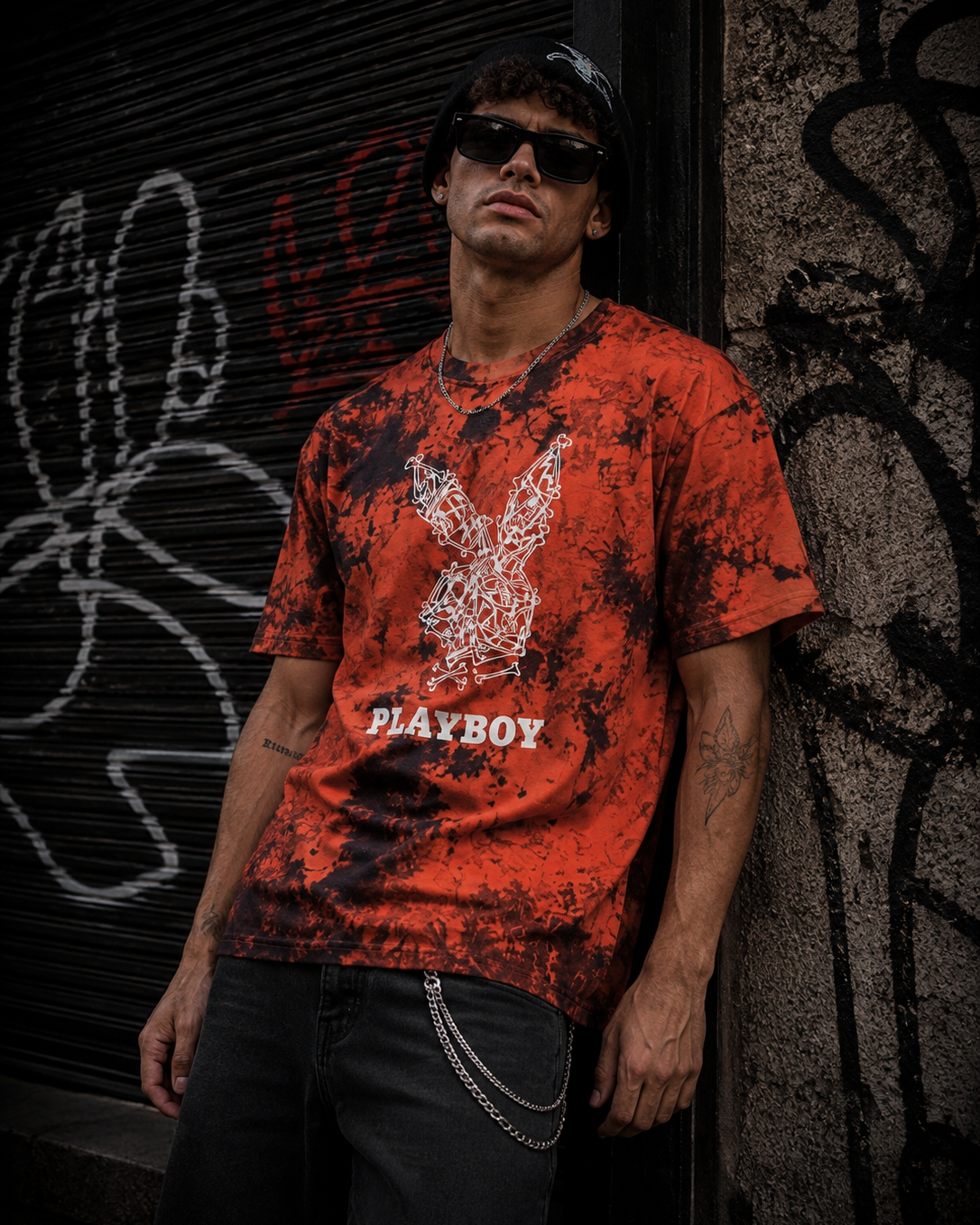 A man wearing a red and black splatter-print t-shirt with a white Playboy bunny logo and the word 'PLAYBOY' printed on the front, posing against a graffiti-covered metal shutter. #Streetwearmen