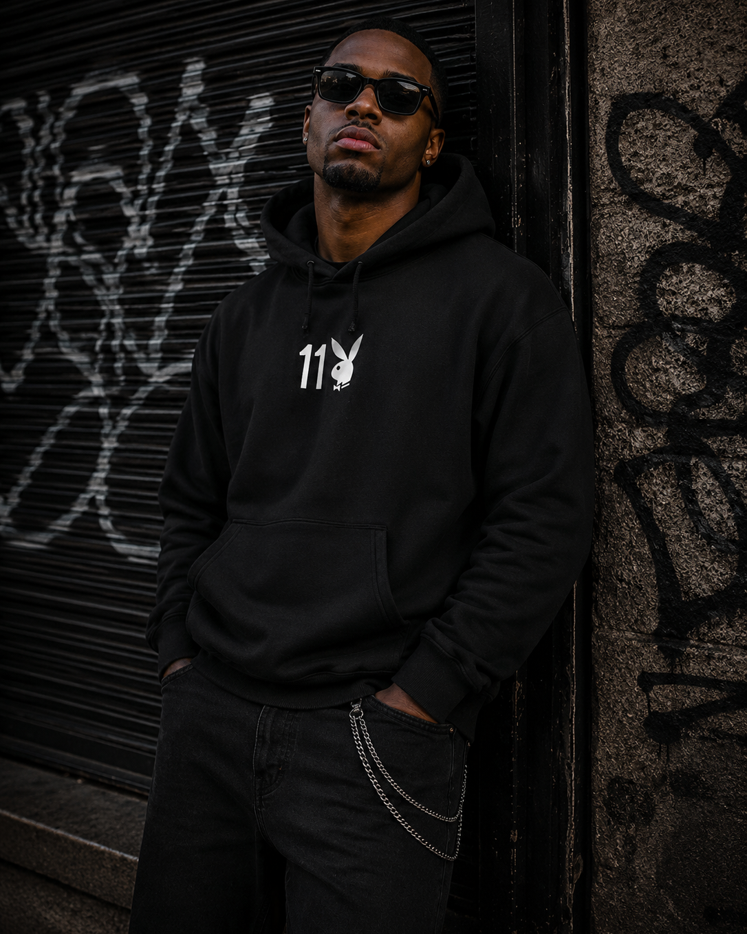 A man wearing a black hoodie with a white Playboy bunny logo and '113' text on the chest, standing against a graffiti-covered metal shutter and concrete wall. #Streetwearmen