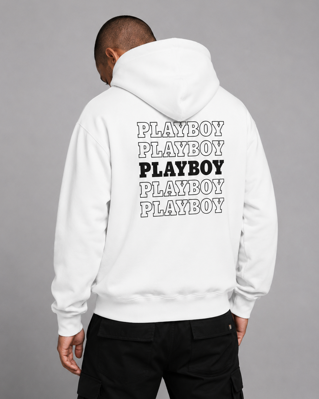 A man viewed from behind wearing a white hoodie with a stacked 'PLAYBOY' graphic printed on the back in black and outlined lettering, paired with black pants, against a solid gray studio background.