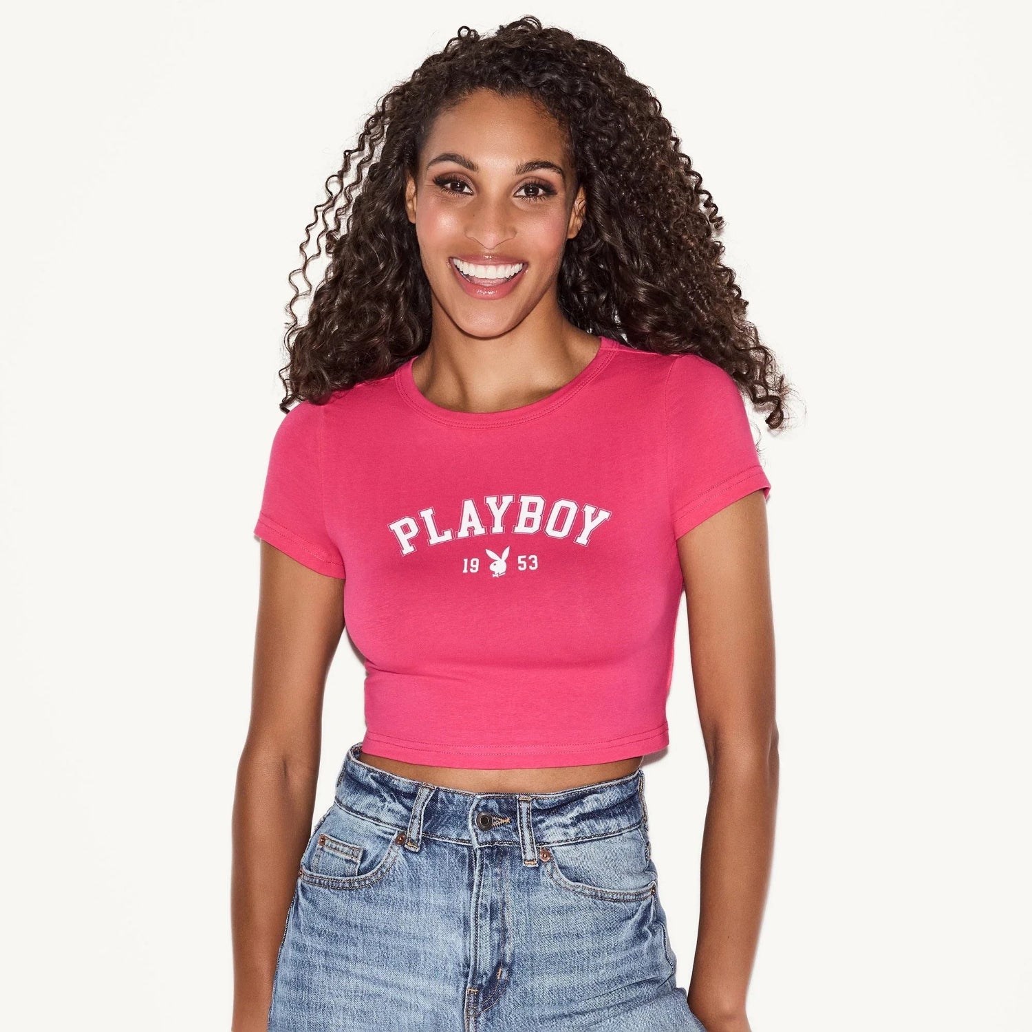 Woman wearing pink cropped baby tee with 'Playboy 1953' graphic and high-waisted jeans
