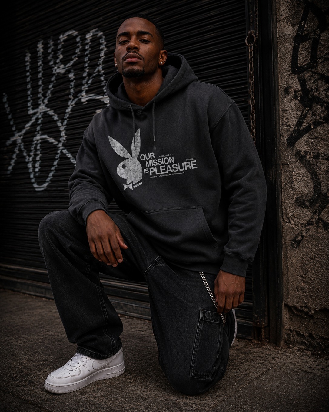 A man kneels in front of a graffiti-covered metal shutter wearing a dark gray hoodie with a white Playboy bunny logo and the text 'OUR MISSION IS PLEASURE' on the front, paired with dark jeans and white sneakers. #Streetwearmen