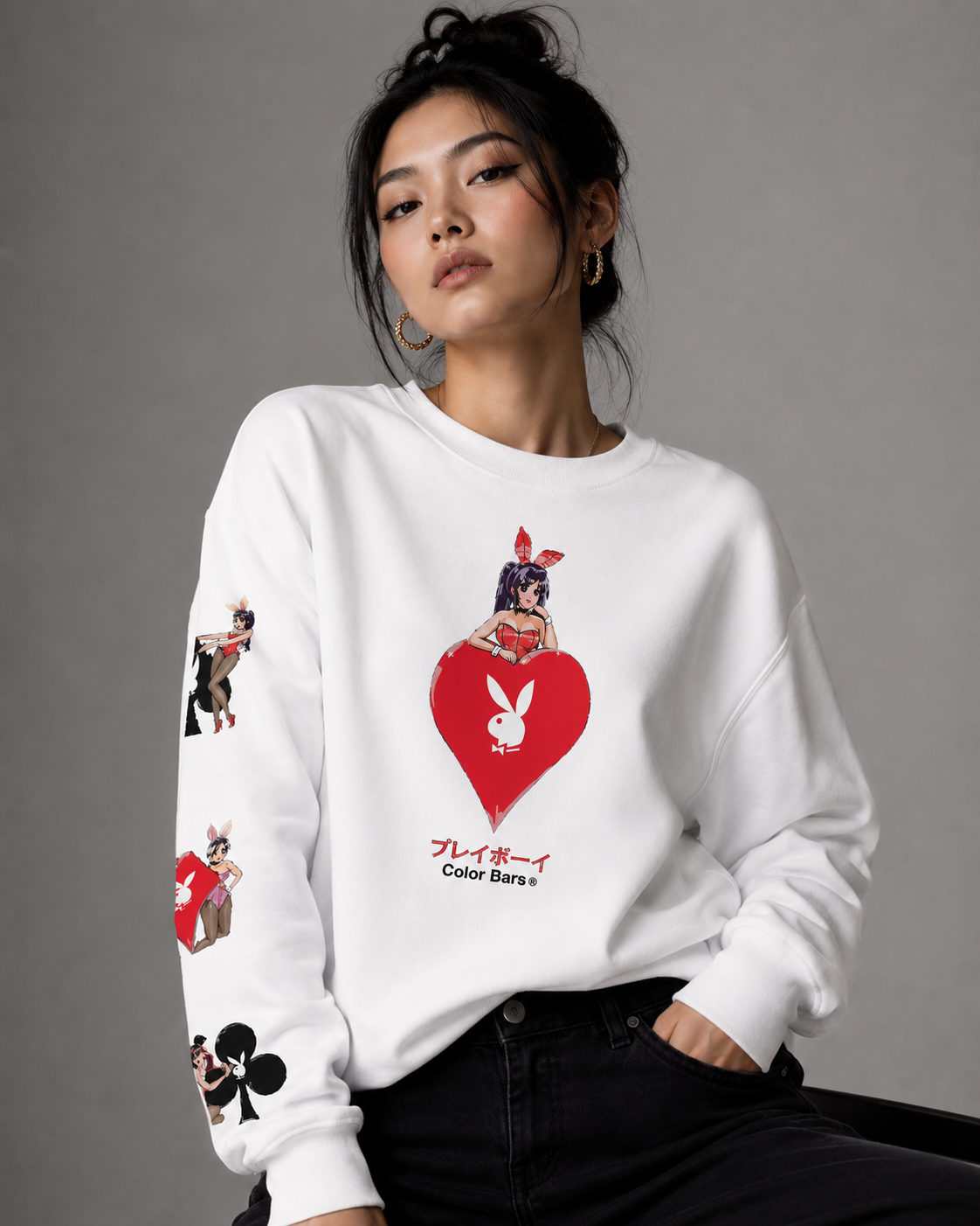 A woman wearing a white long-sleeve sweatshirt with a red heart and rabbit graphic on the front, featuring Playboy branding and illustrations on the sleeves, paired with black jeans against a gray studio background.