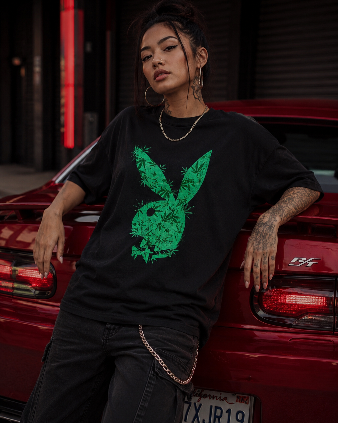 A woman leans against a red car wearing a black oversized T-shirt with a large green bunny graphic on the front, paired with dark jeans and a gold chain necklace. #Streetwearwomen