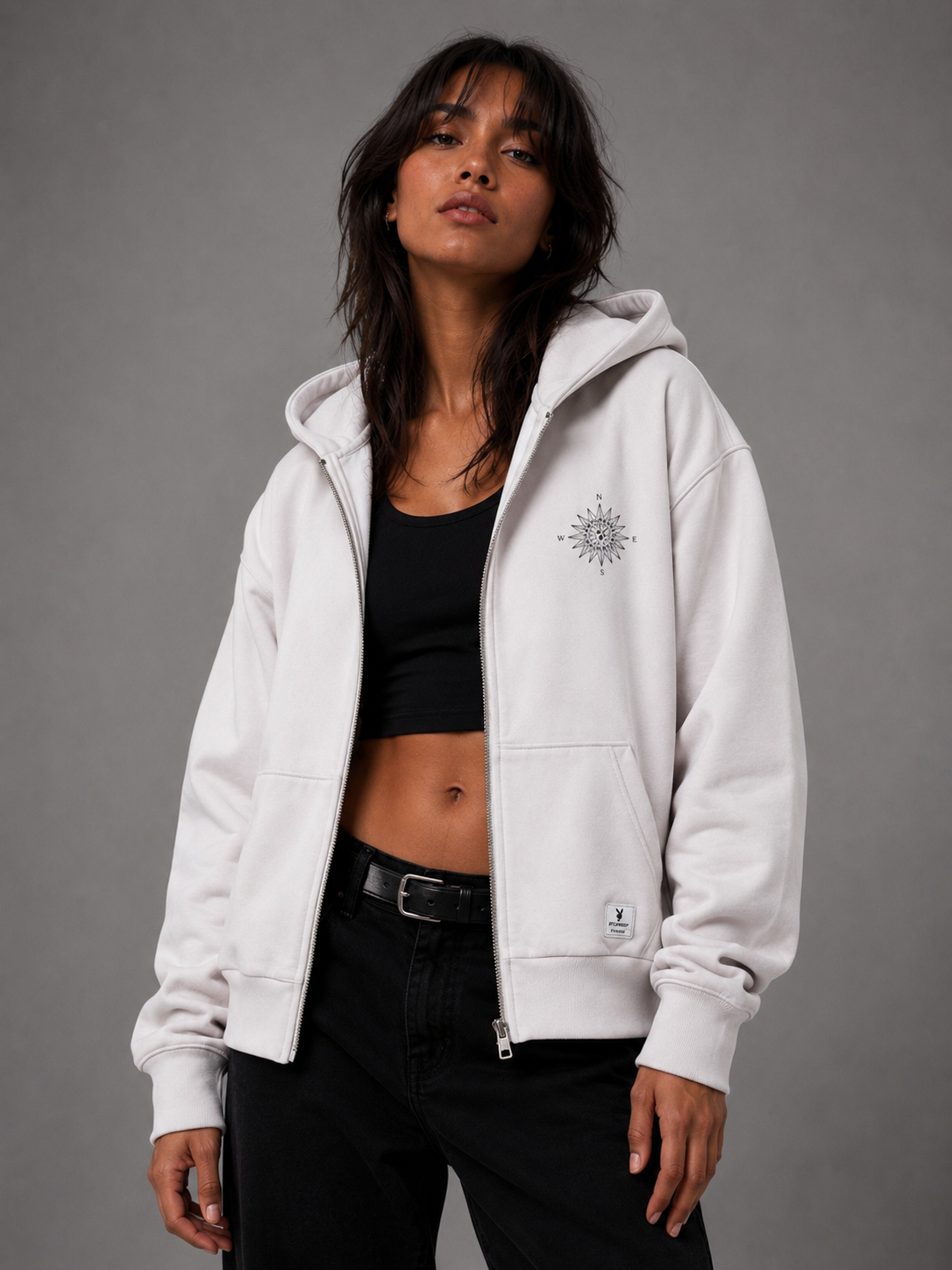 A woman wearing an open white zip-up hoodie with a compass rose graphic over a black crop top, against a solid gray studio background. #Streetwearwomen