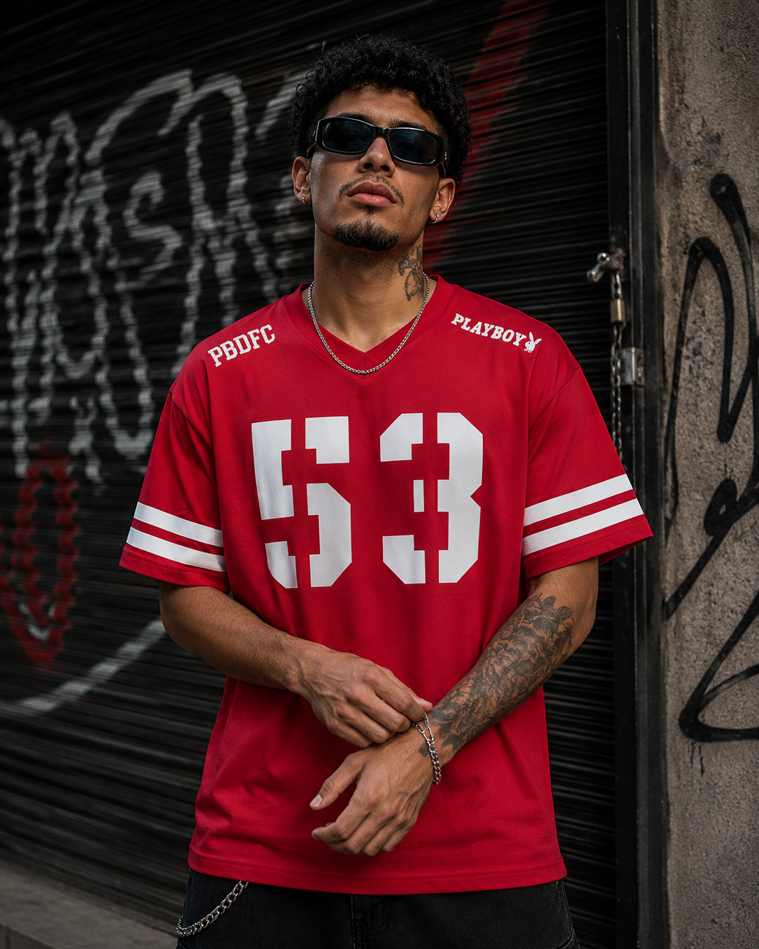 A man wearing a red short-sleeve athletic jersey with white stripes and a large white '53' on the front, standing in front of a graffiti-covered metal shutter. The jersey has 'PBDFC' on the right chest and 'PLAYBOY' with the bunny logo on the left chest. #Streetwearmen